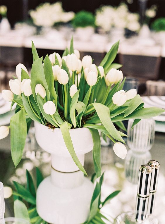 a classy and simple wedding centerpiece of a white vase with white tulips and leaves is a lovely idea for a modern wedding
