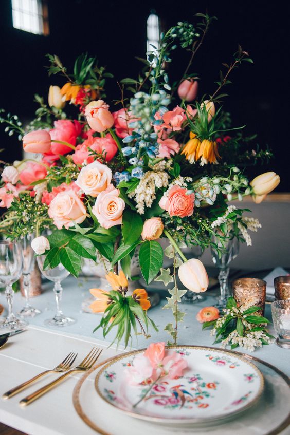 a colorful wedding centerpiece of peachy, pink and hot pink blooms including tulips, blue and white flowers, greenery is amazing