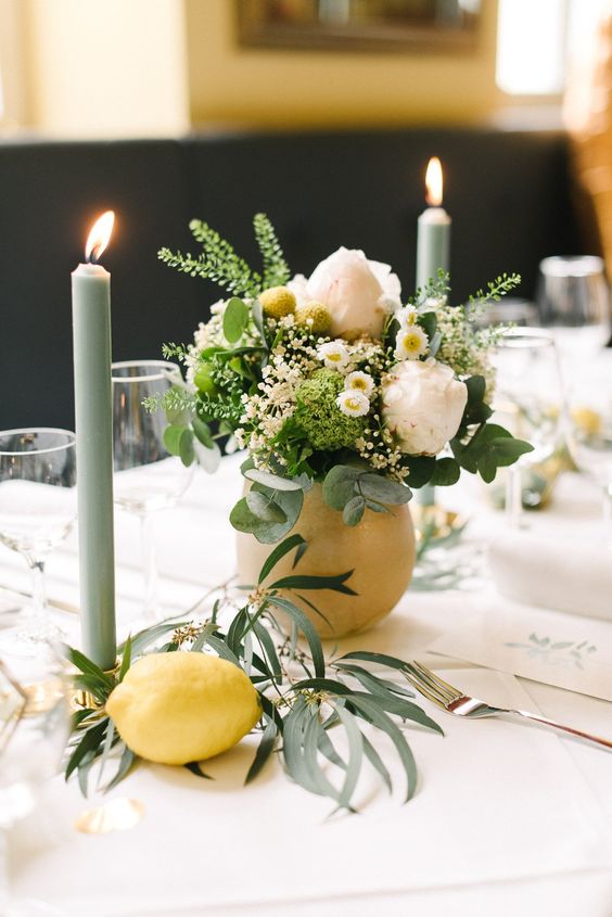 a creative summer wedding centerpiece of a large lemon as a vase for white and yellow blooms and greenery, a lemon and some greenery, olive green candles