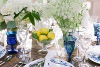 a creative wedding tablescape with an uncovered table, blue floral plates and blue glasses, white blooms and a lemon arrangement in a bowl