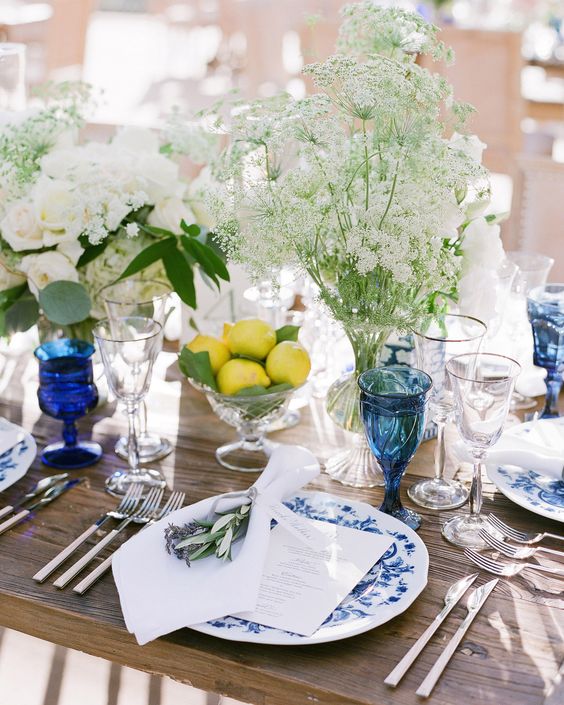a creative wedding tablescape with an uncovered table, blue floral plates and blue glasses, white blooms and a lemon arrangement in a bowl