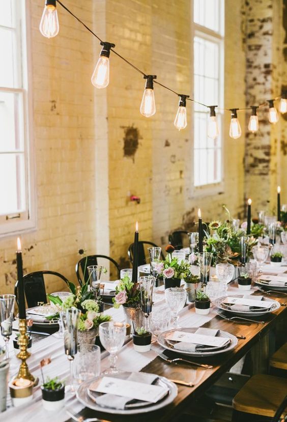 a dramatic wedding reception space with black candles, grey porcelain, greenery and blooms and bulbs over the table