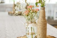 a farmhouse wedding centerpiece of a wood slice, a jar and a bottle wrapped with twine, with baby’s breath and pink roses