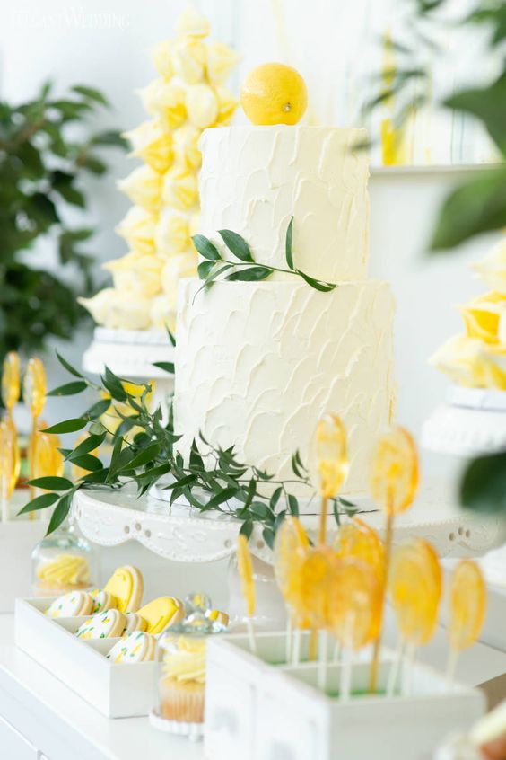 a lemon-themed wedding dessert table with a white buttercream wedding cake with a lemon on top, lemon lollipops and cookies, a lemon meringue cake