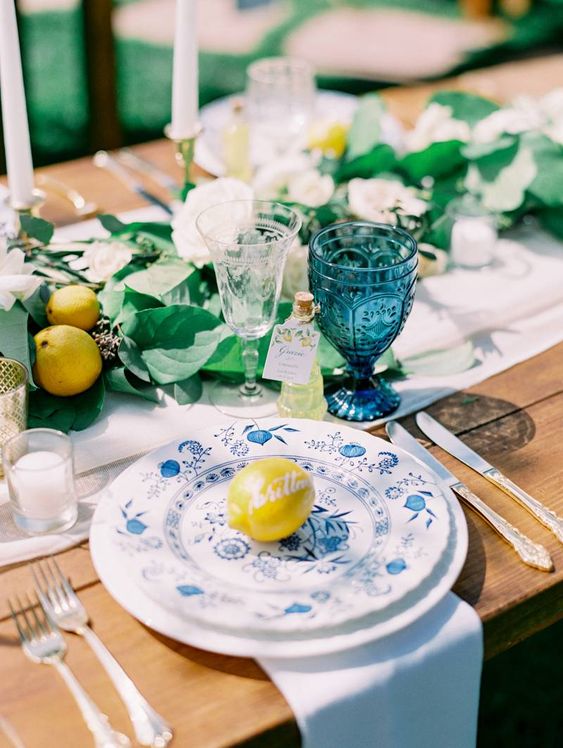 a lovely blue and yellow wedding table setting with a white runner, greenery and lemons, blue printed plates and blue glasses
