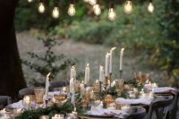 a lovely outdoor wedding reception space with an evergreen runner, candles, ornaments and bulbs over the table is very cozy