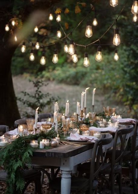 a lovely outdoor wedding reception space with an evergreen runner, candles, ornaments and bulbs over the table is very cozy