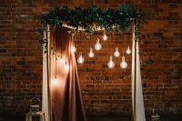 a lovely wedding arch with a copper and a white curtain, greenery and bulbs hanging down plus candles on the floor is a beautiful idea