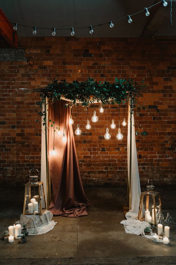 a lovely wedding arch with a copper and a white curtain, greenery and bulbs hanging down plus candles on the floor is a beautiful idea