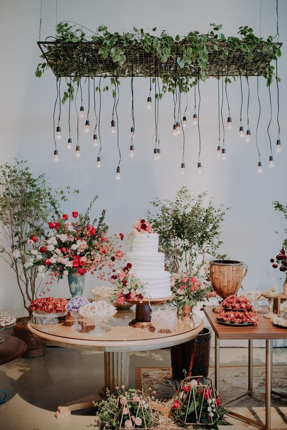 a lovely wedding dessert station with lots of delicious desserts, greenery and bright blooms and a grid with greenery and bulbs hanging down