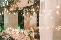 a lush and beautiful wedding reception space with greenery and bulbs over it, lots of candles and blooms on the table and refined vintage chairs