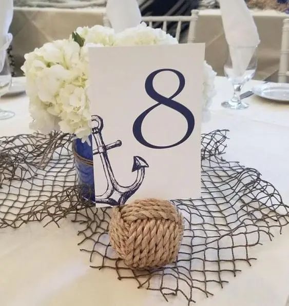 a nautical wedidng centerpiece of net, a rope ball, a table number with an anchor and white blooms in a navy vase