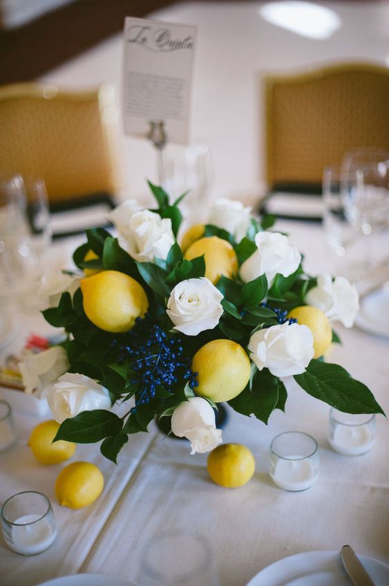 a pretty and simple wedding centerpiece of white roses, lemones, greenery and privet berries plus some candles and lemons around
