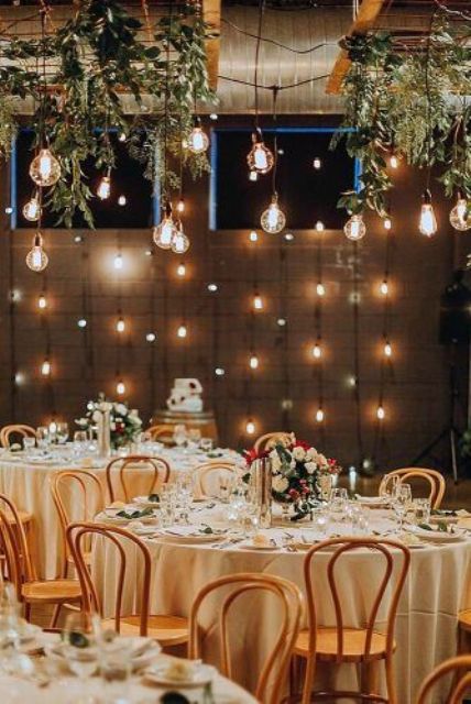 a refined and chic wedding reception space with greenery and bulbs hanging down, a bulb wall and neutral tables with bold blooms