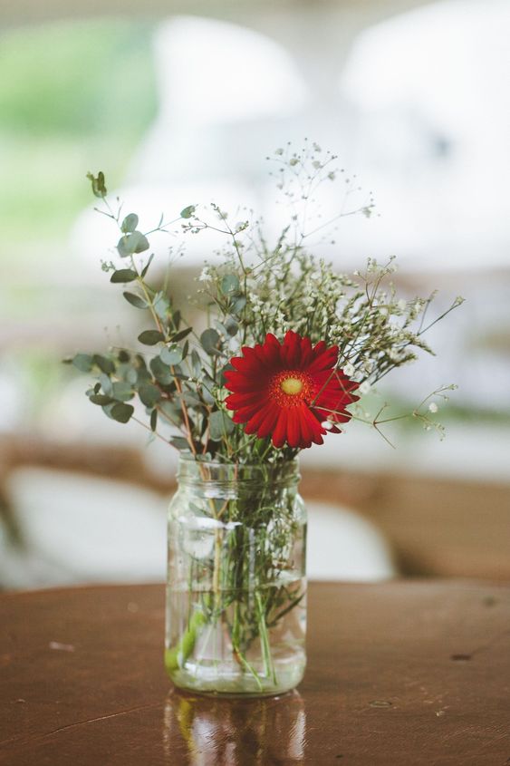a small and simple wedding centerpiece of greenery, baby's breath and a red gerberas in a jar for a rustic wedding