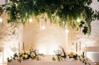 a sophisticated wedding sweetheart table with a greenery and bulb overhead installation and refined blooms on and around the table