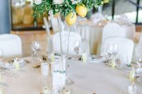 a tall wedding centerpiece of a clear glass vase, white blooms, greenery and some lemons and candles around is a bright and cool idea for a summer wedding