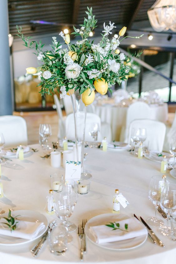 a tall wedding centerpiece of a clear glass vase, white blooms, greenery and some lemons and candles around is a bright and cool idea for a summer wedding