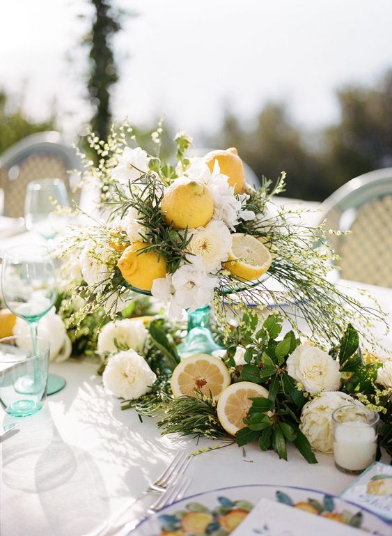 a textural wedding centerpiece of white blooms, cut lemons, candles in glasses and lots of greenery and grasses