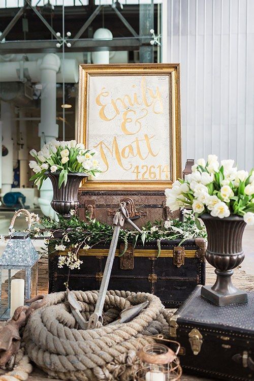 a vintage nautical wedding decor with rope, a large real anchor, candle lanterns, suitcases, white blooms and greenery and a sign