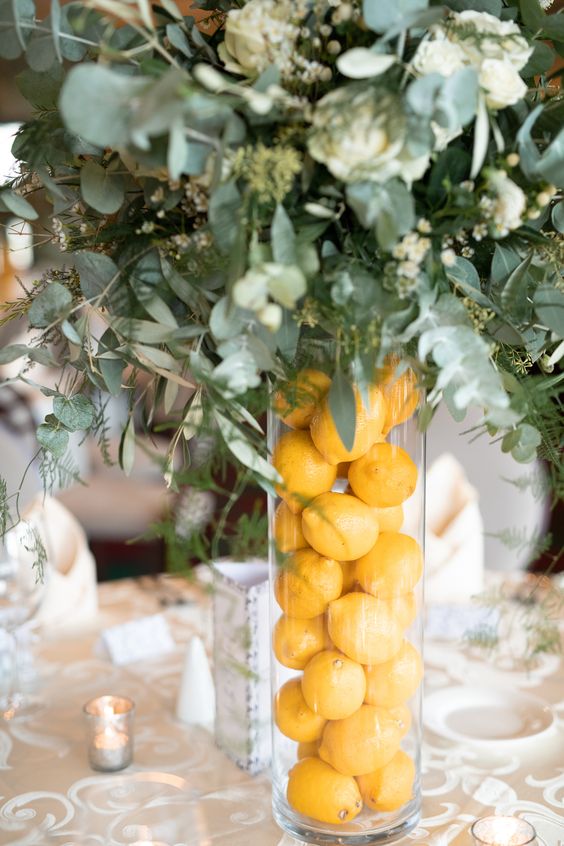 a wedding centerpiece of a tall glass vase filled with lemons and greenery and white blooms is amazing for a bold summer wedding