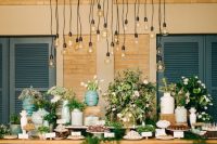 a wedding dessert table with lots of sweets, cakes and various desserts, greeneyr and blooms and some bulbs above to accent it