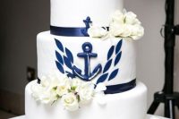 a white and navy wedding cake with a navy anchor, some leaves, white blooms is a stylish idea for a nautical or seaside wedding