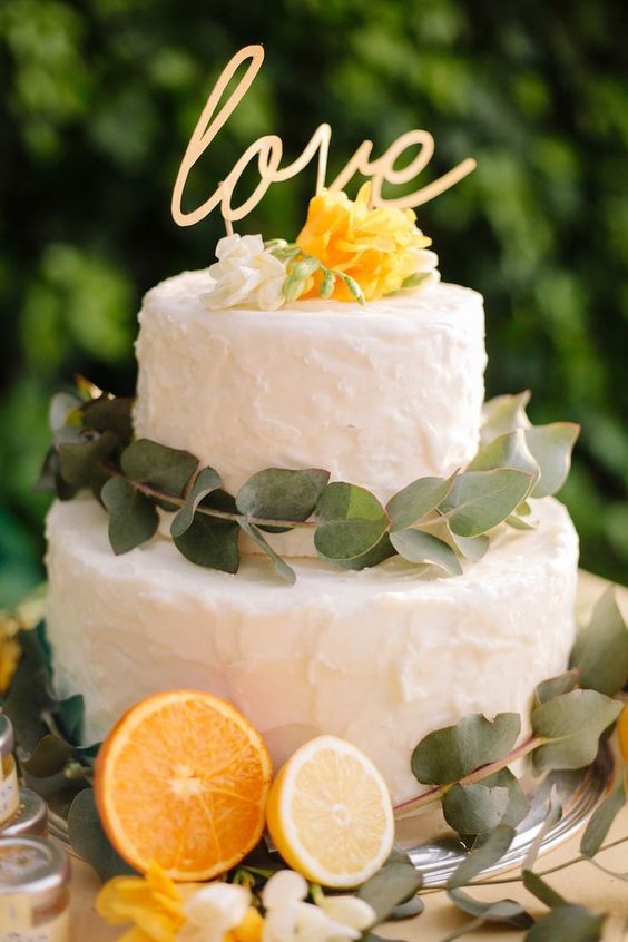 a white textural buttercream decorated with eucalyptus, white and yellow blooms and a calligraphy cake topper