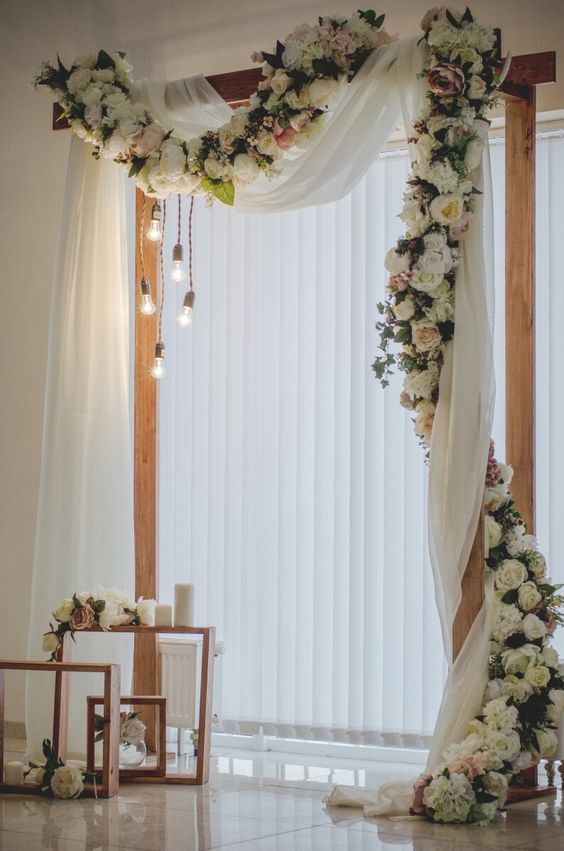 an elegant wedding arch with neutral curtains, white and pink blooms and greenery and bulbs hanging down is amazing