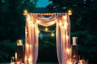 an outdoor wedding arch with neutral curtains, lights and bulbs, pillar candles and petals on the ground is a magical idea