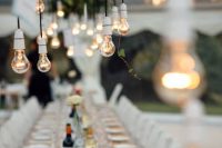an outdoor wedding reception space with neutral and pastel blooms, bulbs hanging down from an overhead greenery installation for a creative reception look
