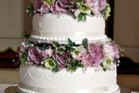 a refined white wedding cake with patterns and lilac and green blooms and greenery between the tiers