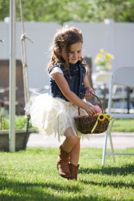 a white top, a neutral tutu skirt, a blue denim waistcoat, cowboy boots and a basket with petals for a fun rustic wedding