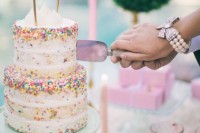 a naked wedding cake with colorful sprinkles, meringues on top and a banner with gold glitter bows