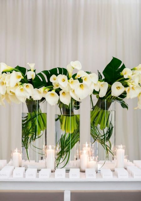 a wedding escort card table with white calla lily arrangements and monstera leaves plus white escort cards are amazing