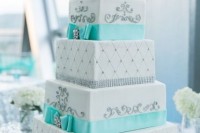a bright turquoise and white square wedding cake with patterns and turquoise ribbons plus a shiny monogram topper