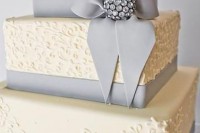 a white square wedding cake with patterns, grey edible ribbons and a large rhinestone piece