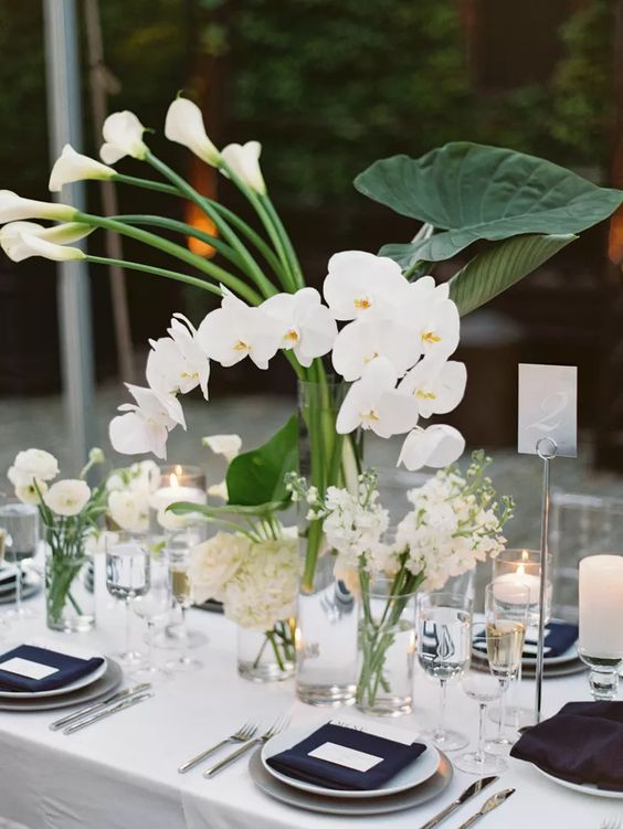 a cluster wedding centerpiece of clear glasses with white callas, orchids, ranunculus and other blooms, all white and chic