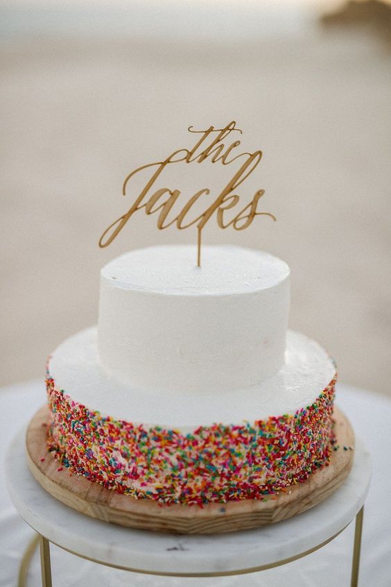 a cool and bold wedding cake with a white and a colorful sprinkle tier and a gold calligraphy topper for a modern wedding