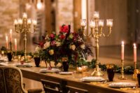 a moody Beauty and the Beast wedding tablescape with candlelabras, gold and burgundy glasses, a lush floral centerpiece and a greenery runner