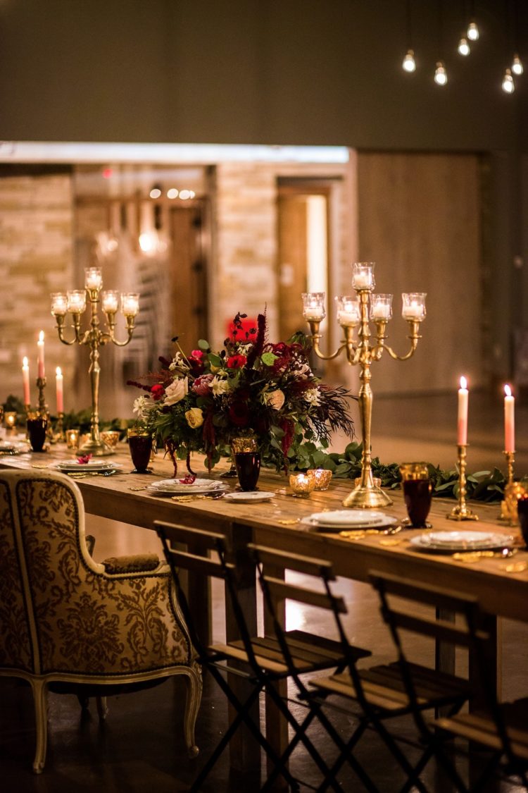 a moody Beauty and the Beast wedding tablescape with candlelabras, gold and burgundy glasses, a lush floral centerpiece and a greenery runner