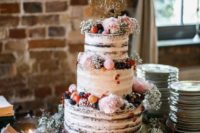 a naked wedding cake topped with berries and fruits, with pink blooms and macarons and an elegant topper