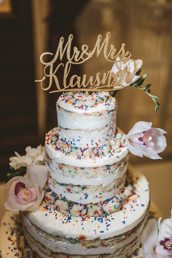 a naked wedding cake with colorful sprinkles, neutral blooms and greenery and a gold calligraphy topper