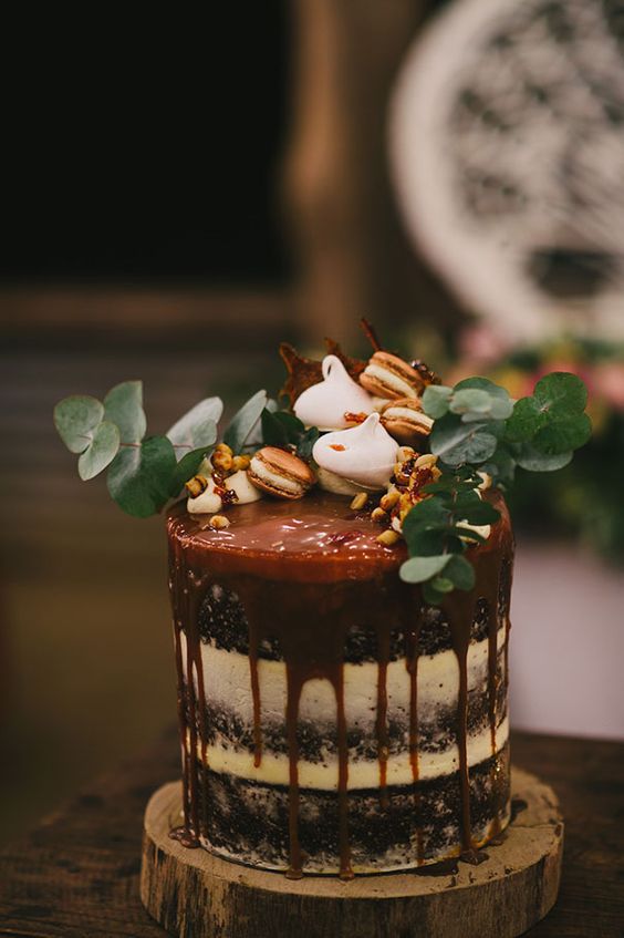 a semi naked wedding cake with caramel drip, topped with eucalyptus, macarons and meringues