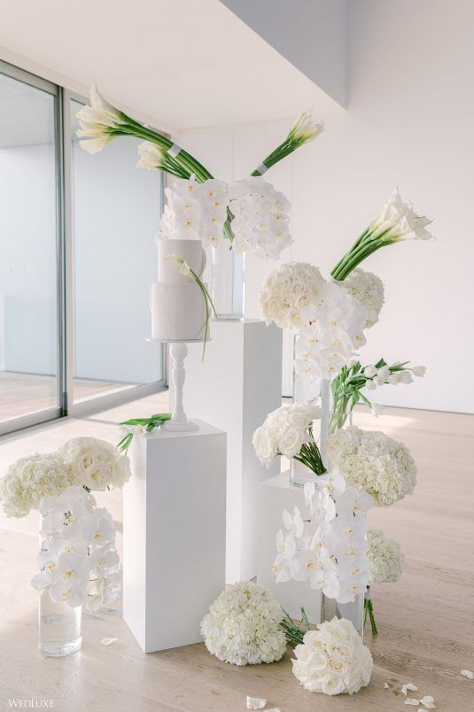 a wedding cake display with white stands, white callas, orchids, roses and hydrangeas and a white wedding cake