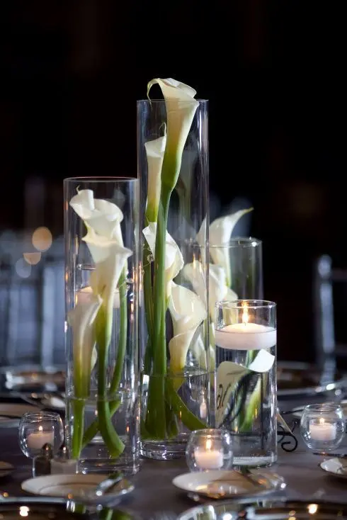 tall glasses with calla lilies and floating candles plus more candles around compose a chic wedding centerpiece