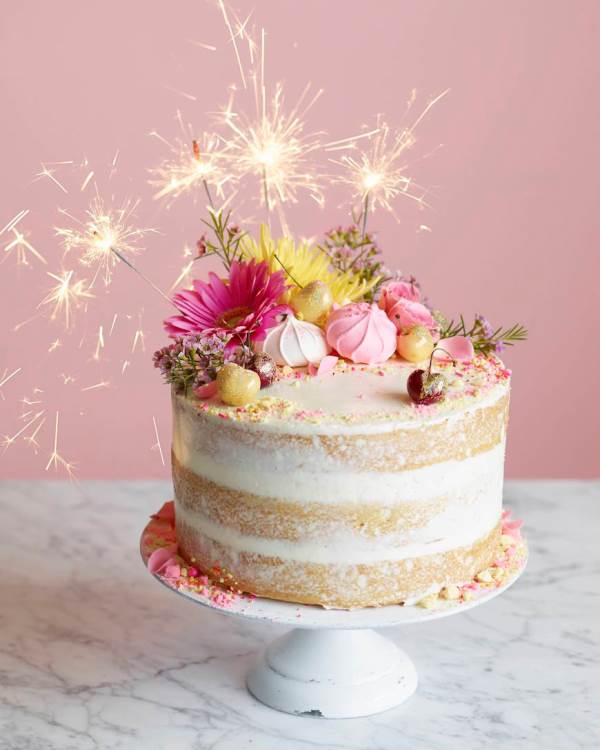 a super bright naked wedding cake with pink and yellow blooms, cherries, sparklers and bright petals and gold leaf