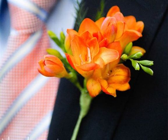 a bold orange freesia wedding boutonniere with greenery is a lovely color accent to the groom's or grooman's look