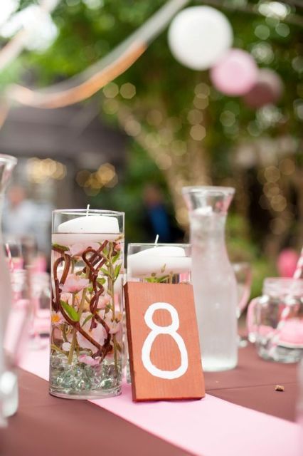 a simple wedding centerpiece of tall glasses with cherry blossom and floating candles plus a table number is a cute idea for a spring wedding