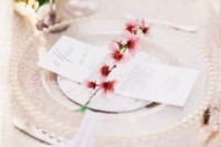 a delicate spring wedding table setting with a printed tablecloth, a clear glass and gold charger, a menu and cherry blossom plus neutral cutlery